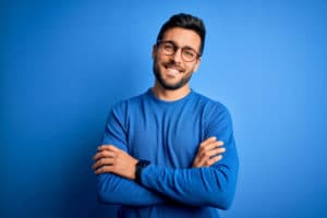 Young handsome man with beard wearing casual sweater and glasses over blue background happy face smiling with crossed arms looking at the camera. Positive person.